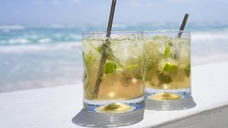 Zwei erfrischende Cocktails mit Limetten und Minze stehen auf einer weißen Holzoberfläche, während der Ozean im Hintergrund glitzert.